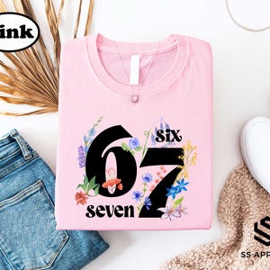 a pink t-shirt with the number "67" and the text "six seven" printed on it, along with a floral design. The t-shirt is displayed on a white background, accompanied by a pair of blue jeans, a pair of white sneakers, and a beige sweater.