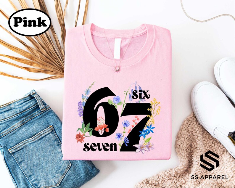 a pink t-shirt with the number "67" and the text "six seven" printed on it, along with a floral design. The t-shirt is displayed on a white background, accompanied by a pair of blue jeans, a pair of white sneakers, and a beige sweater.