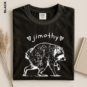 May include: A black t-shirt featuring a white graphic illustration of a raccoon walking. Above the raccoon, the text "jimothy" is printed in a white, handwritten-style font, flanked by two small white heart outlines on either side.