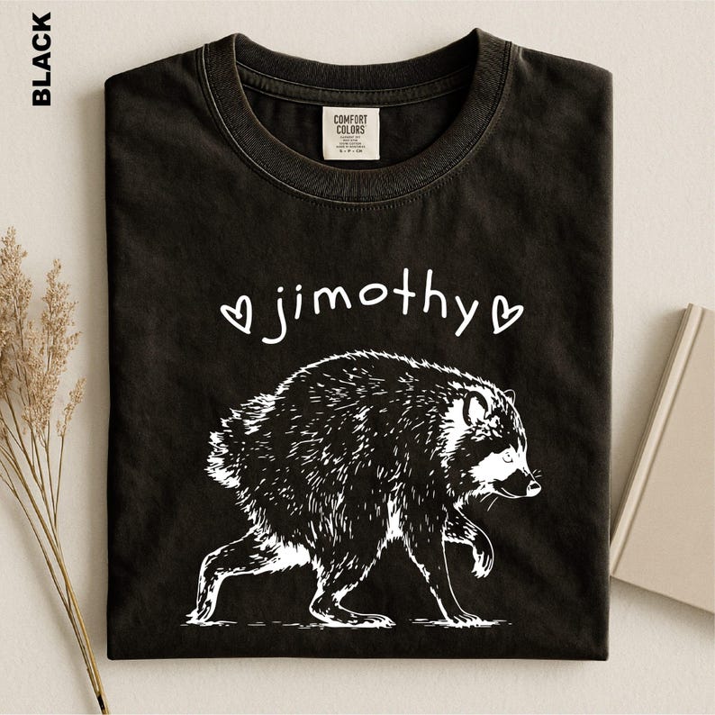 May include: A black t-shirt featuring a white graphic illustration of a raccoon walking. Above the raccoon, the text "jimothy" is printed in a white, handwritten-style font, flanked by two small white heart outlines on either side.