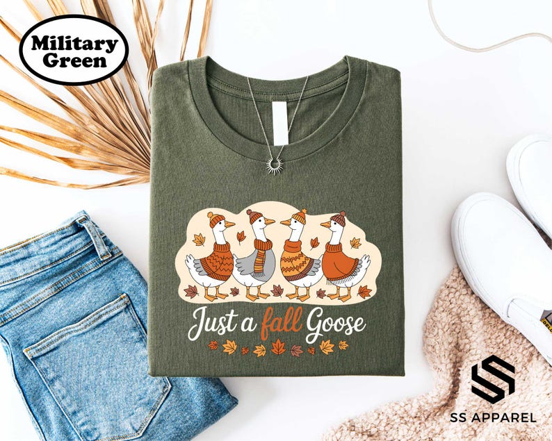 a green t-shirt with a graphic design of four geese wearing orange sweaters and hats, along with the text "Just a fall goose". The t-shirt is displayed on a white background, accompanied by a pair of blue jeans, a pair of white sneakers, and some dried leaves.