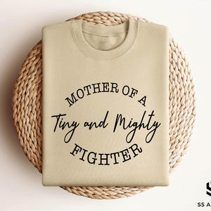 May include: A beige sweatshirt with the text "Mother of a Tiny and Mighty Fighter" printed in black.