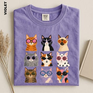 May include: A violet crew neck t-shirt featuring a graphic print of nine different cats wearing various styles of colorful sunglasses. The shirt has a Comfort Colors brand label at the inner neckline and displays the word VIOLET on the side.