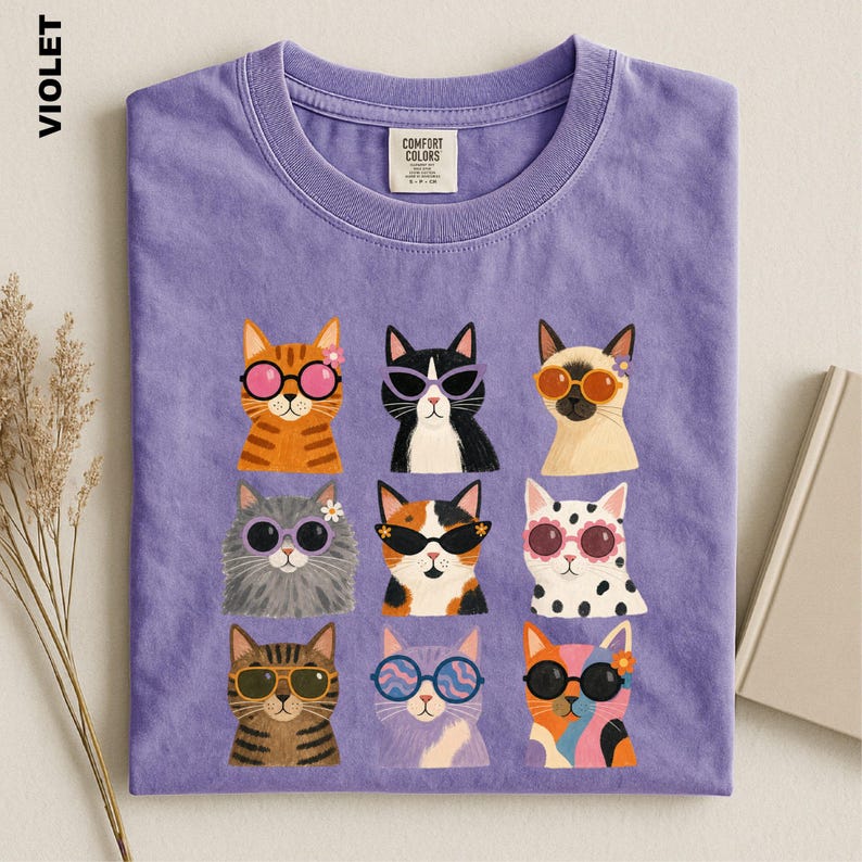 May include: A violet crew neck t-shirt featuring a graphic print of nine different cats wearing various styles of colorful sunglasses. The shirt has a Comfort Colors brand label at the inner neckline and displays the word VIOLET on the side.