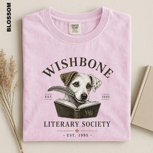 A pink t-shirt with a graphic design featuring a dog holding a book and the text "Wishbone Literary Society" printed on it.