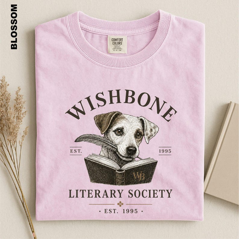 A pink t-shirt with a graphic design featuring a dog holding a book and the text "Wishbone Literary Society" printed on it.