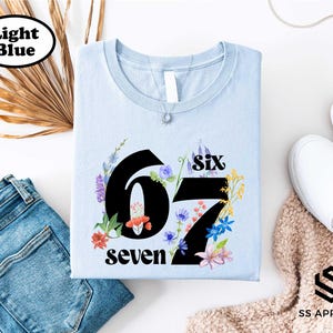 a light blue t-shirt with the number "67" and the text "seven" printed on it, along with a floral design. The t-shirt is placed on a white background, and there are some other clothing items and accessories visible in the image.
