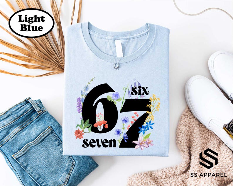 a light blue t-shirt with the number "67" and the text "seven" printed on it, along with a floral design. The t-shirt is placed on a white background, and there are some other clothing items and accessories visible in the image.
