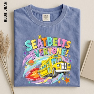 May include: A blue jean coloured t-shirt featuring a graphic of a yellow school bus flying through space with a rocket flame. The text reads SEATBELTS EVERYONE! in colourful letters. The design includes planets, stars, and a cartoon character.
