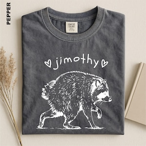 May include: A pepper-colored t-shirt featuring a white graphic of a raccoon walking. Above the raccoon, the text reads "jimothy" in a handwritten font, flanked by two small white heart outlines. The shirt has a crew neckline and a label.