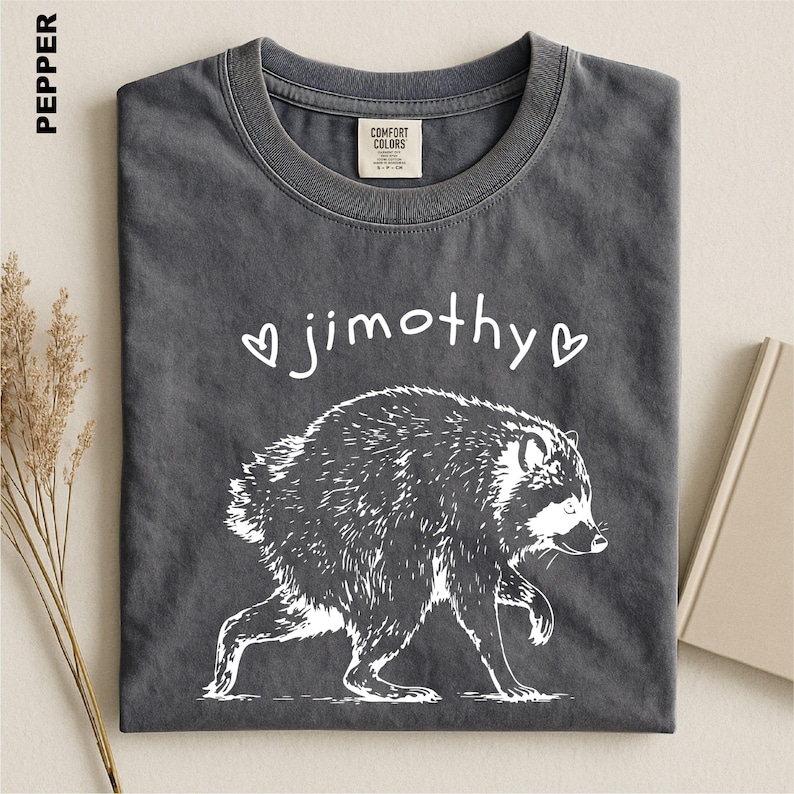 May include: A pepper-colored t-shirt featuring a white graphic of a raccoon walking. Above the raccoon, the text reads "jimothy" in a handwritten font, flanked by two small white heart outlines. The shirt has a crew neckline and a label.