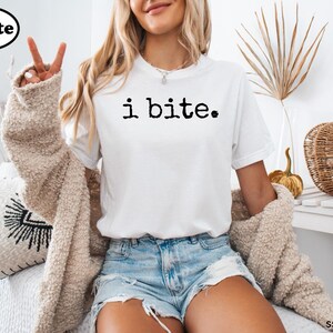 A woman wearing a white t-shirt with the text "i bite" and denim shorts is sitting on a couch, making a peace sign with her fingers.