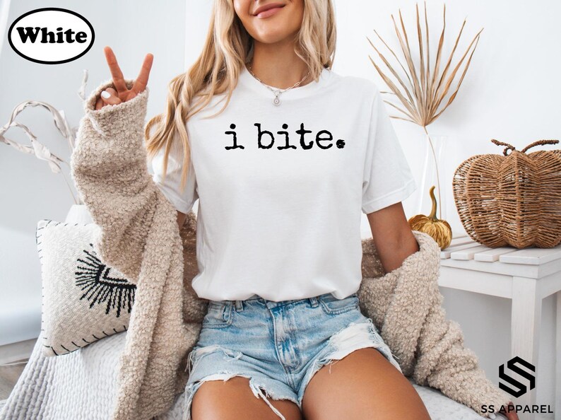 A woman wearing a white t-shirt with the text "i bite" and denim shorts is sitting on a couch, making a peace sign with her fingers.