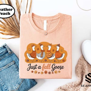 a pink t-shirt with a graphic design of four ducks wearing winter clothing, accompanied by the text "Just a fall Goose". The t-shirt is displayed alongside a pair of jeans, a pair of white sneakers, and a beige sweater.