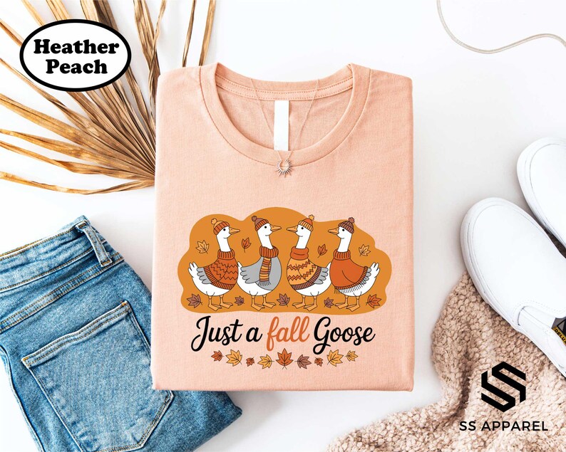 a pink t-shirt with a graphic design of four ducks wearing winter clothing, accompanied by the text "Just a fall Goose". The t-shirt is displayed alongside a pair of jeans, a pair of white sneakers, and a beige sweater.