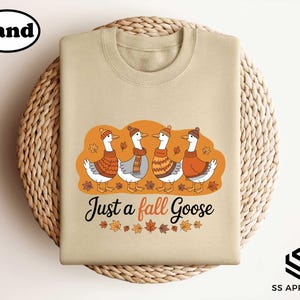 A beige t-shirt with a graphic design featuring four ducks wearing orange and brown sweaters, surrounded by autumn leaves, and the text "Just a fall Goose" below the image.