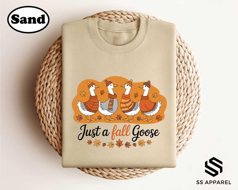 A beige t-shirt with a graphic design featuring four ducks wearing orange and brown sweaters, surrounded by autumn leaves, and the text "Just a fall Goose" below the image.