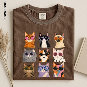 May include: A brown short-sleeve t-shirt featuring a graphic print of nine cats wearing various colorful sunglasses and small flowers. The shirt has a crew neckline and a Comfort Colors brand label on the inside collar. The word ESPRESSO is printed vertically on the left side.