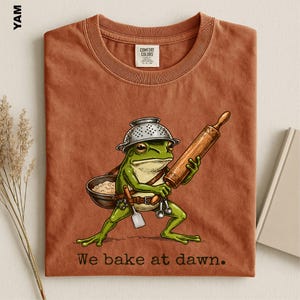 Op de afbeelding: Een roestkleurig t-shirt met een afbeelding van een groene kikker die een vergiet als hoed draagt, een houten deegroller vasthoudt en een kom deeg draagt. De tekst "We bake at dawn." staat eronder gedrukt in een donker schreeflettertype.