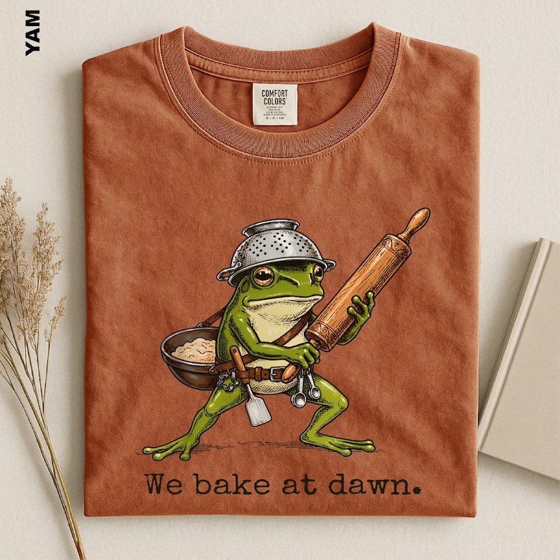 Op de afbeelding: Een roestkleurig t-shirt met een afbeelding van een groene kikker die een vergiet als hoed draagt, een houten deegroller vasthoudt en een kom deeg draagt. De tekst "We bake at dawn." staat eronder gedrukt in een donker schreeflettertype.