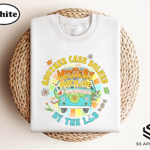 May include: White crew neck sweatshirt featuring a colorful graphic of a teal van with the text "Mystery Machine Diagnostics" and the phrase "Another Case Solved By The Lab". The design includes science-themed illustrations.