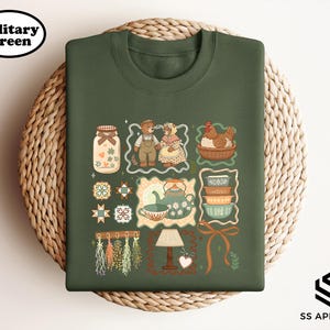 May include: Military green sweatshirt with a whimsical illustration featuring a jar, teddy bears, a chicken in a basket, teacups, and other decorative elements. The sweatshirt has a crew neck and a soft, comfortable appearance. The text "SS APPAREL" is in the bottom right corner.