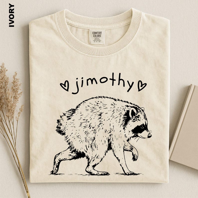 May include: An ivory t-shirt featuring a black line art illustration of a raccoon walking. Above the raccoon, the text "jimothy" is printed in a lowercase, handwritten-style font, flanked by two small black heart symbols on either side.