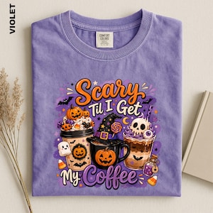 May include: A violet crew neck t-shirt featuring a graphic of three coffee cups with Halloween themes, including pumpkins, a skull, and a witch hat. The text reads Scary Til I Get My Coffee. The shirt has a Comfort Colors brand neck label.