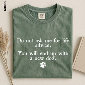 May include: A moss green crew neck t-shirt featuring the white printed text: Do not ask me for life advice. You will end up with a new dog. A white paw print icon is positioned below the text. The shirt has a Comfort Colors brand neck label.