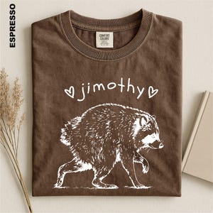 May include: An espresso-colored t-shirt featuring a white graphic of a raccoon walking. Above the raccoon, the text reads "jimothy" in a handwritten font, flanked by two small heart outlines. The shirt has a crew neckline and a Comfort Colors label.