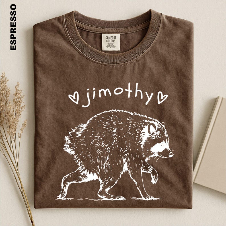 May include: An espresso-colored t-shirt featuring a white graphic of a raccoon walking. Above the raccoon, the text reads "jimothy" in a handwritten font, flanked by two small heart outlines. The shirt has a crew neckline and a Comfort Colors label.