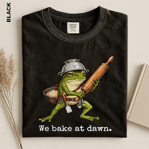 Op de afbeelding: Een zwart t-shirt met een afbeelding van een groene kikker die een vergiet als helm draagt, een houten deegroller vasthoudt en een kom deeg draagt. De kikker heeft een riem met keukengerei. De tekst luidt, We bake at dawn.
