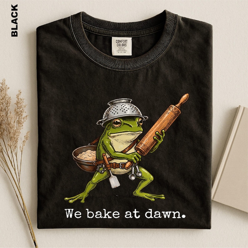 Op de afbeelding: Een zwart t-shirt met een afbeelding van een groene kikker die een vergiet als helm draagt, een houten deegroller vasthoudt en een kom deeg draagt. De kikker heeft een riem met keukengerei. De tekst luidt, We bake at dawn.