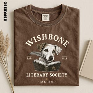 a brown t-shirt with a graphic design depicting a dog holding a book, with text that reads "Wishbone Literary Society" and "EST. 1995".