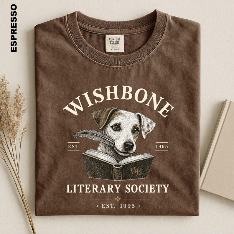 a brown t-shirt with a graphic design depicting a dog holding a book, with text that reads "Wishbone Literary Society" and "EST. 1995".