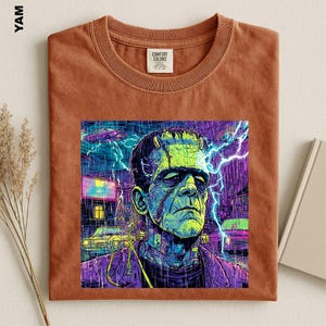 May include: A rust-colored crew neck t-shirt featuring a graphic print of a green-skinned monster with bolts in its neck. The artwork depicts a rainy night scene with bright blue lightning, a yellow car, and a purple building in the background.