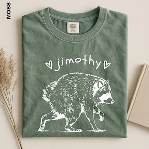 May include: A moss green crew neck t-shirt featuring a white graphic of a raccoon and the text "jimothy" flanked by two small hearts. The shirt has a Comfort Colors brand label on the inside of the collar and a short-sleeved design.