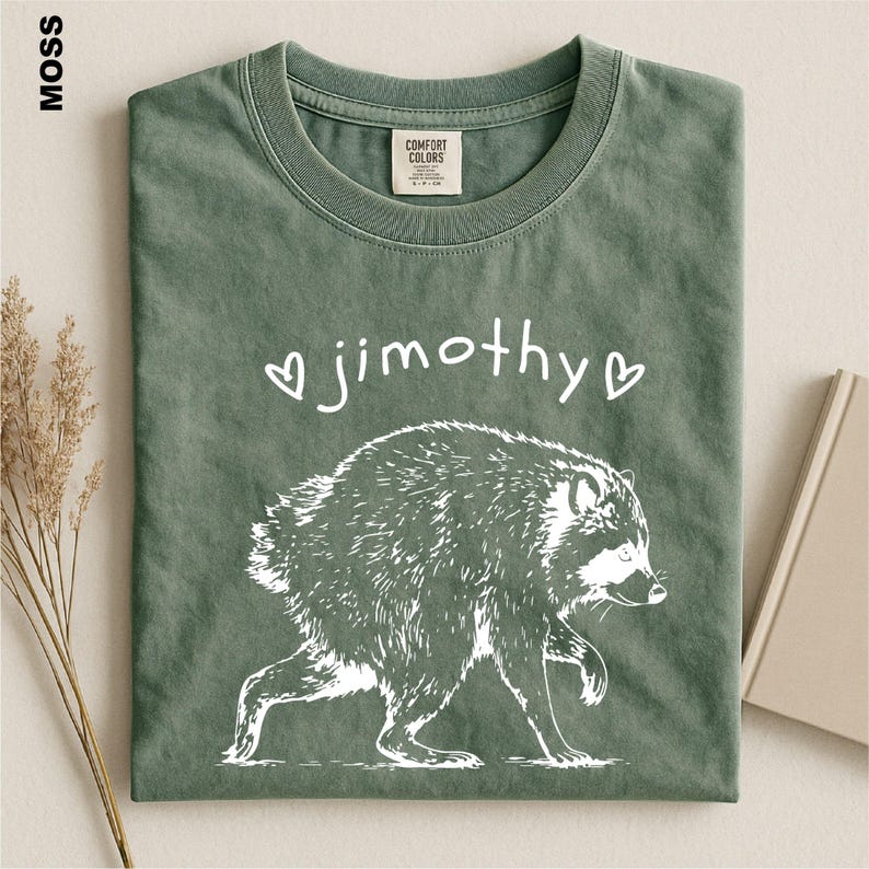 May include: A moss green crew neck t-shirt featuring a white graphic of a raccoon and the text "jimothy" flanked by two small hearts. The shirt has a Comfort Colors brand label on the inside of the collar and a short-sleeved design.