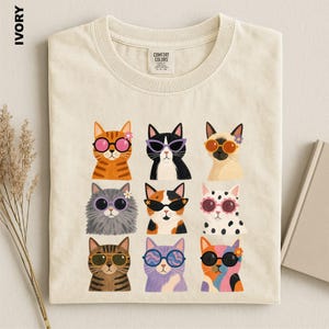 May include: An ivory t-shirt featuring a graphic print of nine cats arranged in a grid. Each cat wears colorful sunglasses and some have small flowers on their heads. The shirt has a crew neckline and a Comfort Colors brand label at the neck.