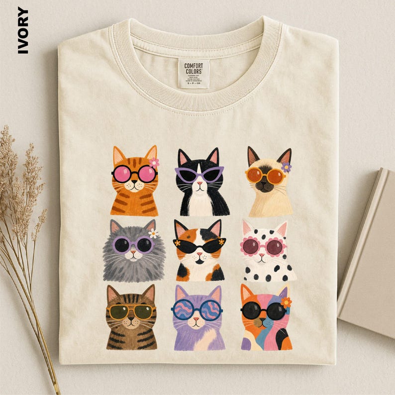 May include: An ivory t-shirt featuring a graphic print of nine cats arranged in a grid. Each cat wears colorful sunglasses and some have small flowers on their heads. The shirt has a crew neckline and a Comfort Colors brand label at the neck.
