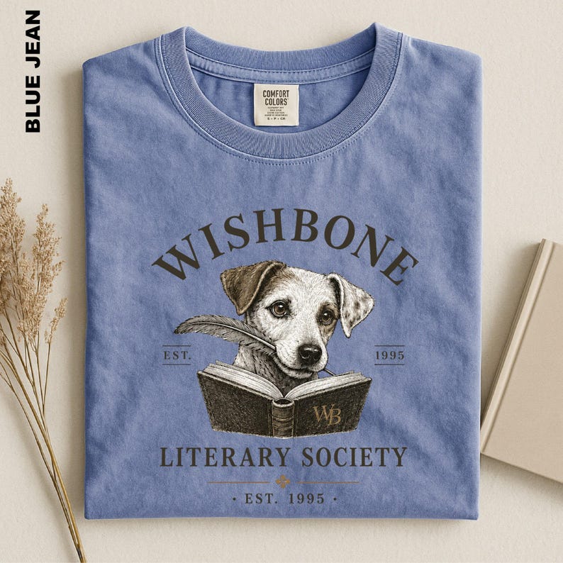 A blue t-shirt with a graphic design featuring a dog holding a book, along with text that reads "Wishbone Literary Society" and "EST. 1995".