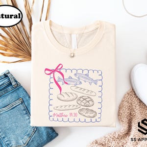 a casual outfit consisting of a white t-shirt with a graphic design, a pair of blue jeans, white sneakers, and a beige sweater.