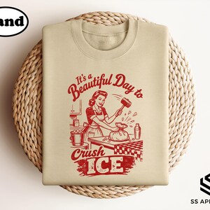 May include: Sand-colored sweatshirt with a retro graphic in red. The graphic features a woman crushing ice with a mallet, accompanied by the text "It's a Beautiful Day to Crush ICE."