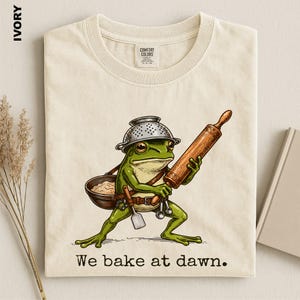 Op de afbeelding: Een ivoorkleurig t-shirt met een afbeelding van een groene kikker die een metalen vergiet als helm draagt, een houten deegroller vasthoudt en een kom en keukengerei draagt. De tekst "We bake at dawn." staat in het zwart onder de afbeelding.