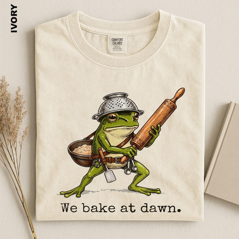 Op de afbeelding: Een ivoorkleurig t-shirt met een afbeelding van een groene kikker die een metalen vergiet als helm draagt, een houten deegroller vasthoudt en een kom en keukengerei draagt. De tekst "We bake at dawn." staat in het zwart onder de afbeelding.