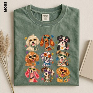 Puede incluir: Una camiseta de cuello redondo color verde musgo con un estampado gr&aacute;fico de nueve razas de perros diferentes que llevan gafas de sol coloridas y pa&ntilde;uelos estampados, decorado con peque&ntilde;as formas de flores y estrellas. Etiqueta Comfort Colors.