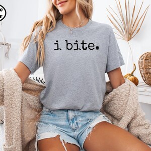 A woman wearing a gray t-shirt with the text "i bite" printed on it, paired with denim shorts and a beige jacket. She is sitting in a chair and has a basket nearby.