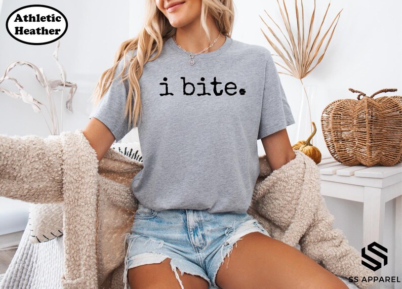 A woman wearing a gray t-shirt with the text "i bite" printed on it, paired with denim shorts and a beige jacket. She is sitting in a chair and has a basket nearby.