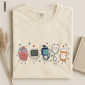 May include: An ivory t-shirt featuring a graphic print of five anthropomorphic medical items: a heart, a heart rate monitor, a blood pressure cuff, a stethoscope, and a pill bottle, all with smiling faces, surrounded by small colourful stars and hearts.