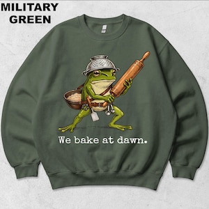 Op de afbeelding: Een militair groen sweatshirt met ronde hals en een afbeelding van een groene kikker die een vergiet als helm draagt, een houten deegroller vasthoudt en een kom en spatel bij zich heeft. De tekst "We bake at dawn." staat in het wit onder de kikker.